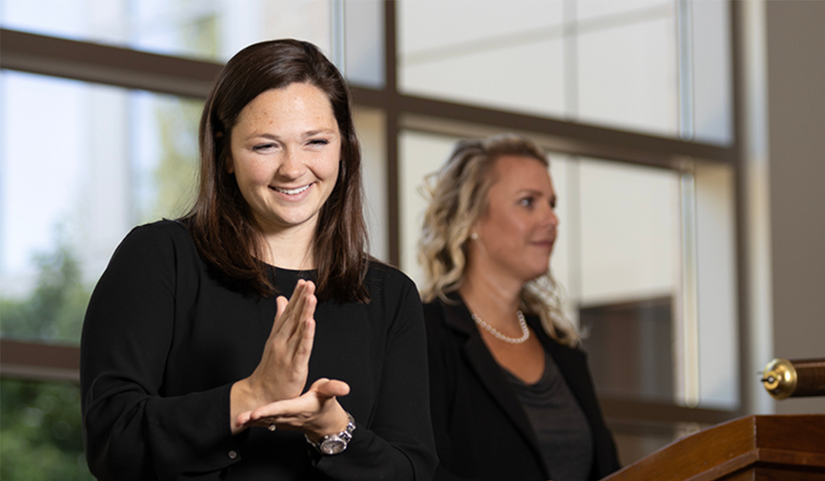 A woman in a black long-sleeve top smiling while providing sign language interpretation next to a speaker at a podium.