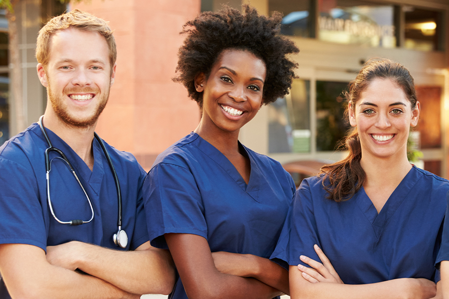 Three smiling healthcare professionals in navy scrubs standing together outside a hospital building, one wearing a stethoscope.