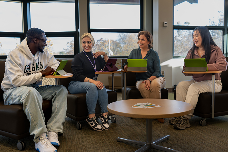 Four people sit in a circle in a sunlit lounge area, smiling and talking while using tablets on swivel desks attached to their chairs. One person wears a white "Cougars" hoodie, and large windows in the background look out onto trees.
