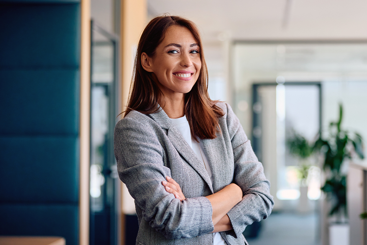A smiling professional woman in a gray blazer stands with arms crossed in a bright office hallway.