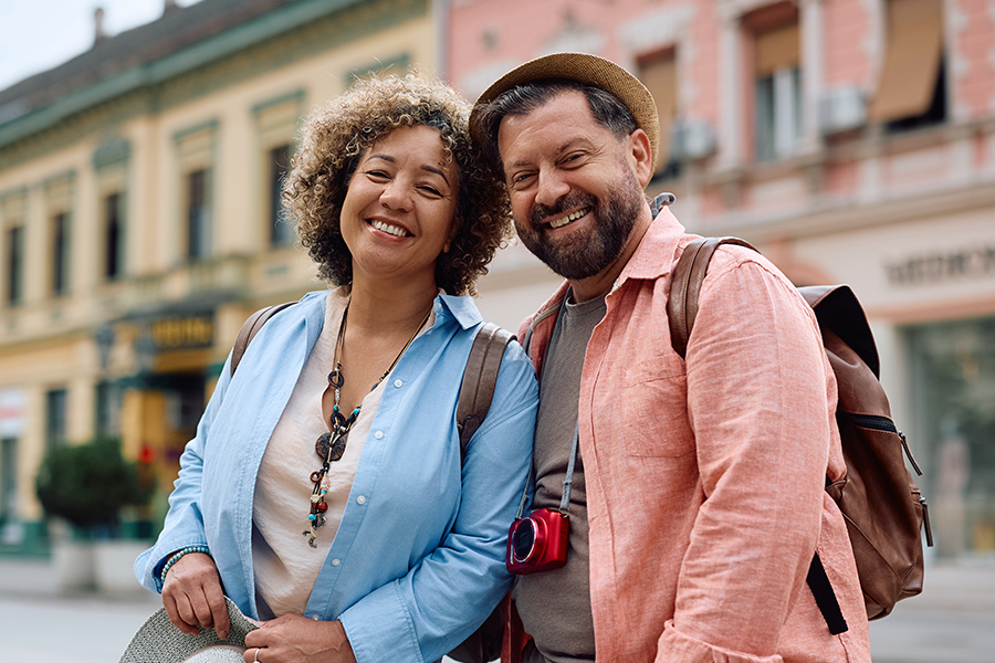 A smiling couple stand close together on a city street, wearing backpacks and casual travel clothing, with colorful historic buildings behind them.