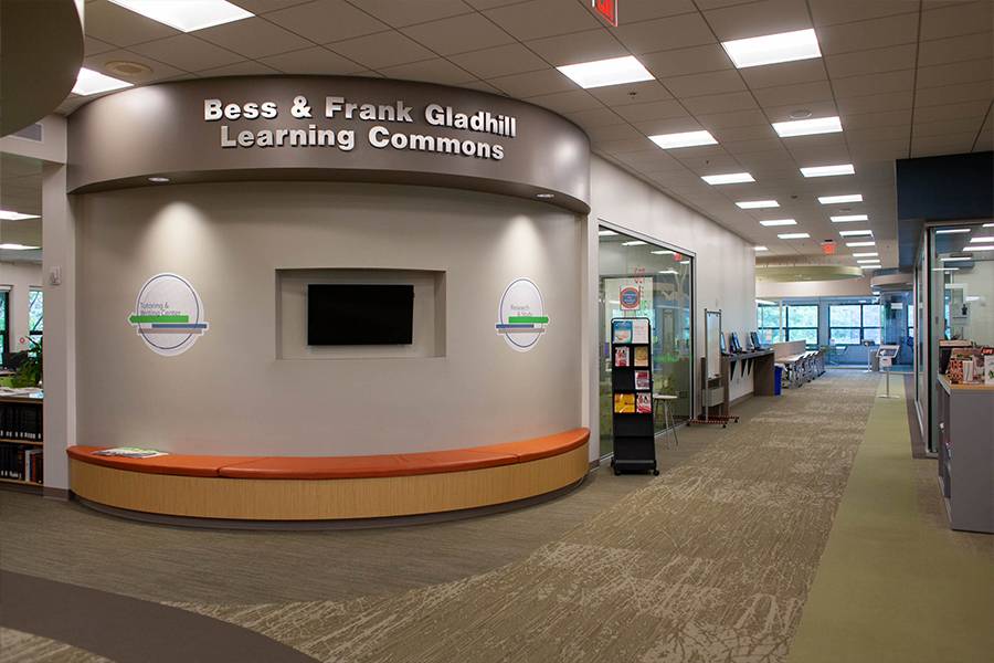 The entrance to the Bess & Frank Gladhill Learning Commons. A curved, grey accent wall features the facility's name in raised silver lettering above a long, orange cushioned bench. To the right, a wide carpeted hallway leads past glass-walled study rooms and informational kiosks into a bright, open academic space.