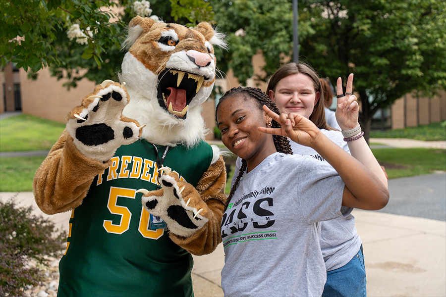 Two smiling students posing outdoors on a campus quad with Freddie the Cougar, the college mascot. One student wears a grey PASS program t-shirt and both students are making peace signs.