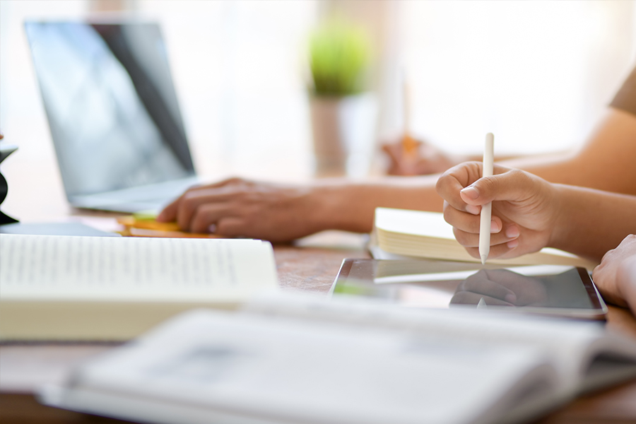A bright, close-up shot of a student’s hand using a white stylus on a tablet. Open textbooks and another person working on a laptop are visible in the soft-focus foreground and background, suggesting a collaborative study environment.