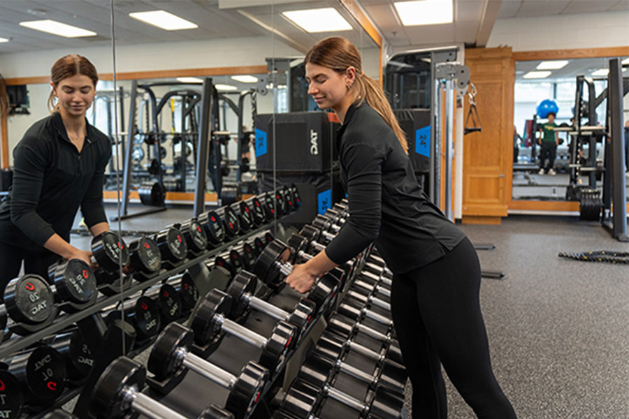 A student in a black athletic top selects a heavy dumbbell from a multi-tier rack in a bright, modern strength and conditioning lab; large mirrors and professional power racks are visible in the background.