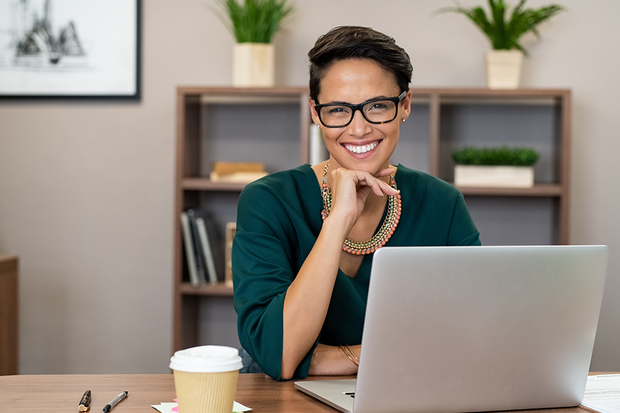 A smiling woman with short hair and glasses seated at a desk with a laptop and coffee cup in a bright, plant-filled office.