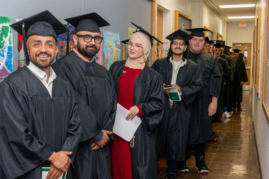 Diverse group of graduates in black caps and gowns smiling in a hallway during a graduation ceremony.
