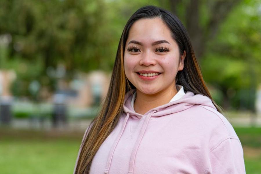 A young woman with long dark hair and blonde highlights smiling outdoors in a light pink hoodie.