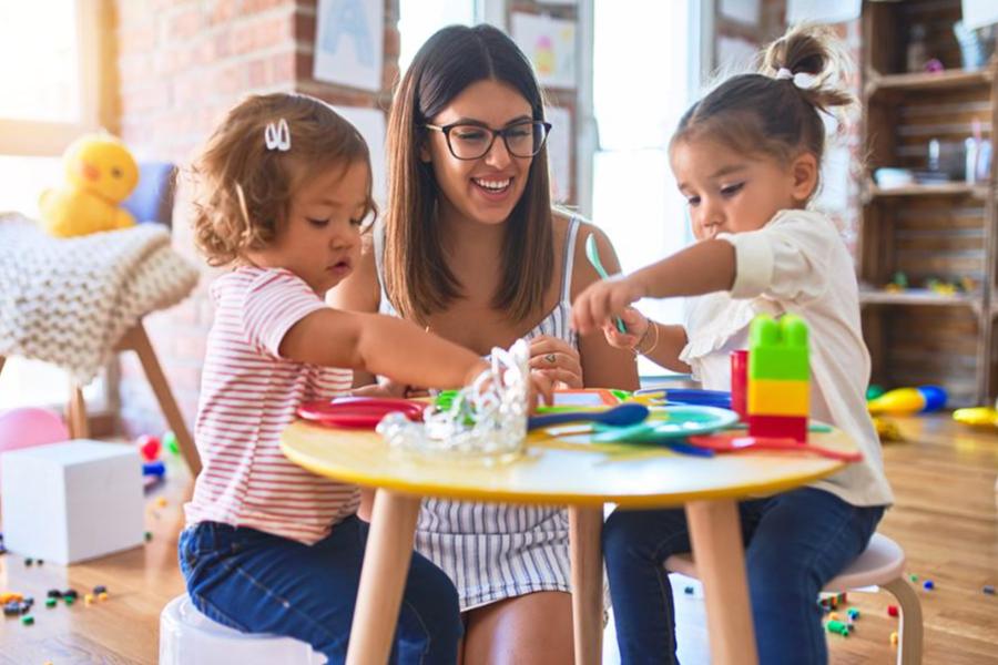 A smiling teacher or daycare provider playing with two young children at a small table in a bright classroom.