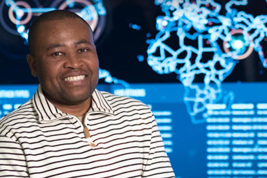 A smiling man in a striped quarter-zip sweater stands in front of a digital world map display in a cybersecurity lab.