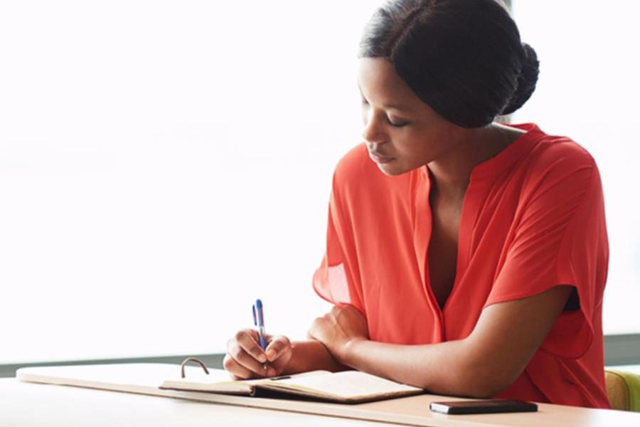 A focused woman in a bright orange top writing in a journal or notebook at a desk.