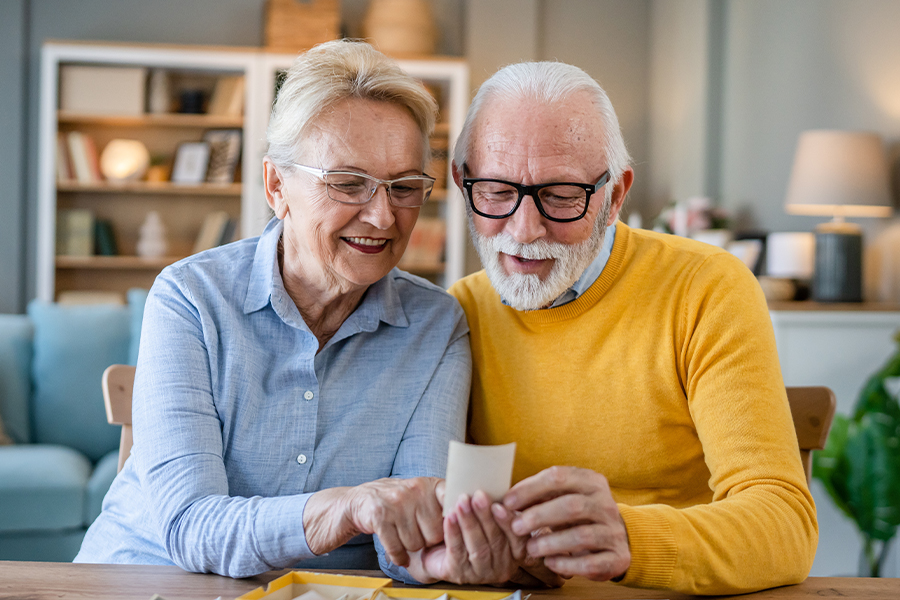 Two smiling older adults wearing glasses sitting together at a table, looking at a photograph and sharing a warm moment at home.