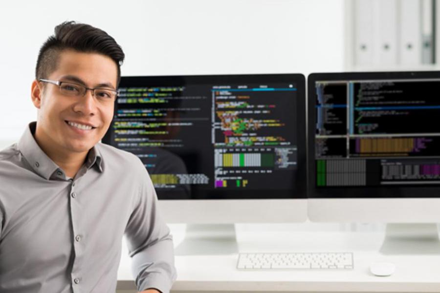 A smiling man with glasses sits in front of two monitors displaying computer code in a bright office.