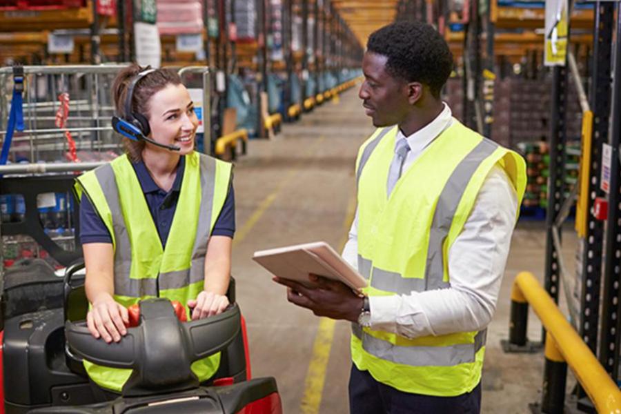 Two warehouse workers in yellow high-visibility vests converse in a large distribution center, with one seated on a forklift wearing a headset and the other standing and holding a tablet, with long rows of shelving visible in the background.