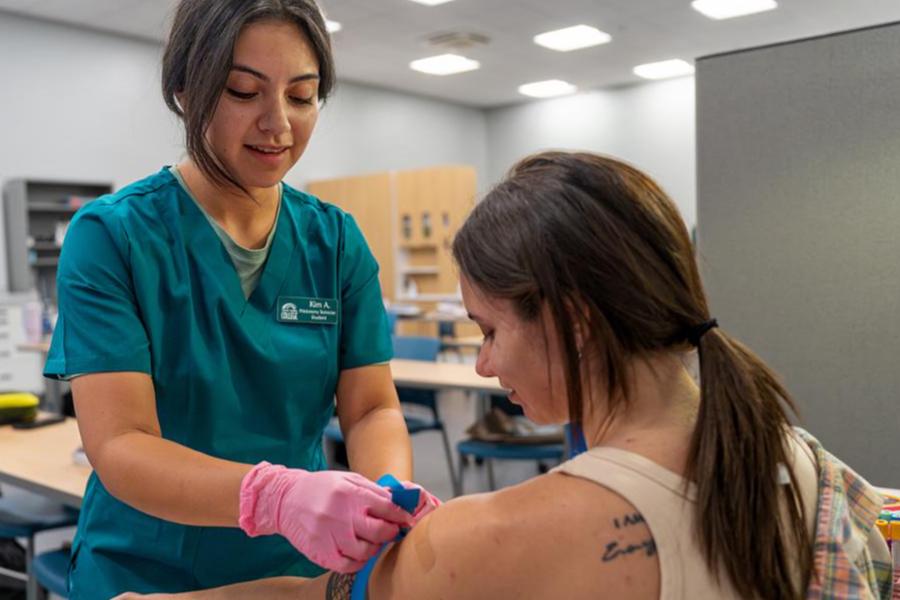FCC phlebotomy student in teal scrubs and pink gloves preparing to draw blood from a patient's arm in a clinical training classroom