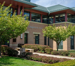 The exterior of Linganore Hall, a brick building with a green roof on the Frederick Community College campus, surrounded by trees and a manicured lawn on a sunny day.