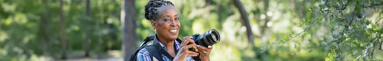 A smiling older woman with silver braided hair holds a DSLR camera while outdoors in a lush green wooded area, wearing a vest and carrying a backpack.