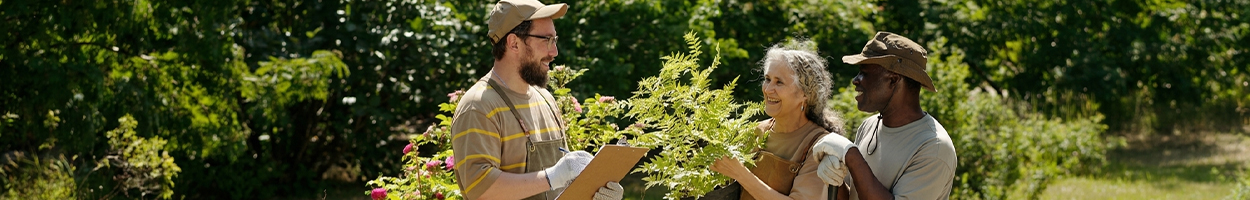 Three adults smile and interact outdoors in a lush garden setting, with one man holding a clipboard and another person holding a leafy plant while wearing gardening gloves.