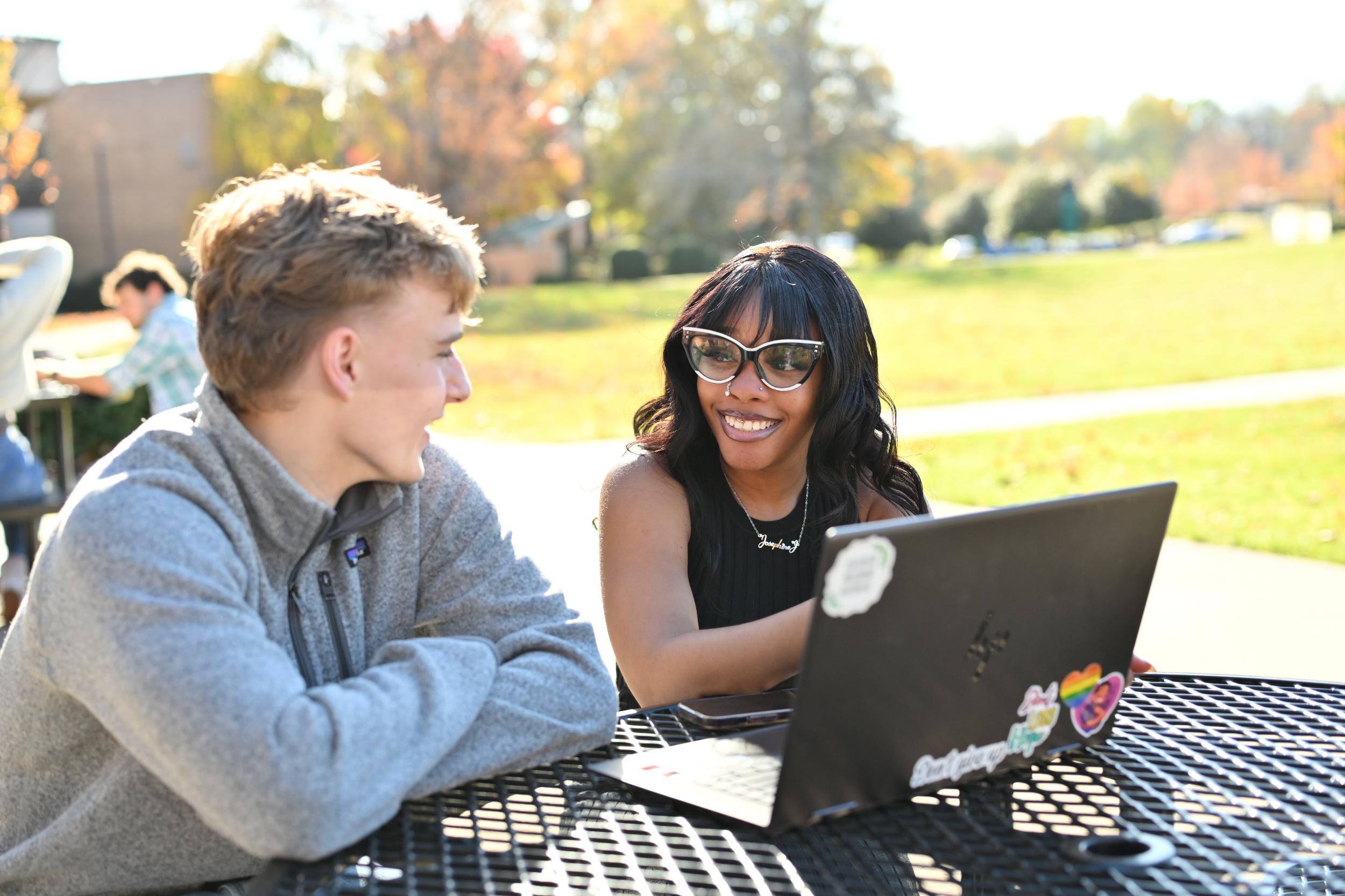 2 students sitting at outside table. Woman has open laptop. Man has grey hoodie