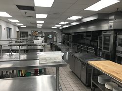 A professional commercial kitchen and culinary lab featuring stainless steel prep tables, industrial ovens, ranges with ventilation hoods, and tiled flooring.