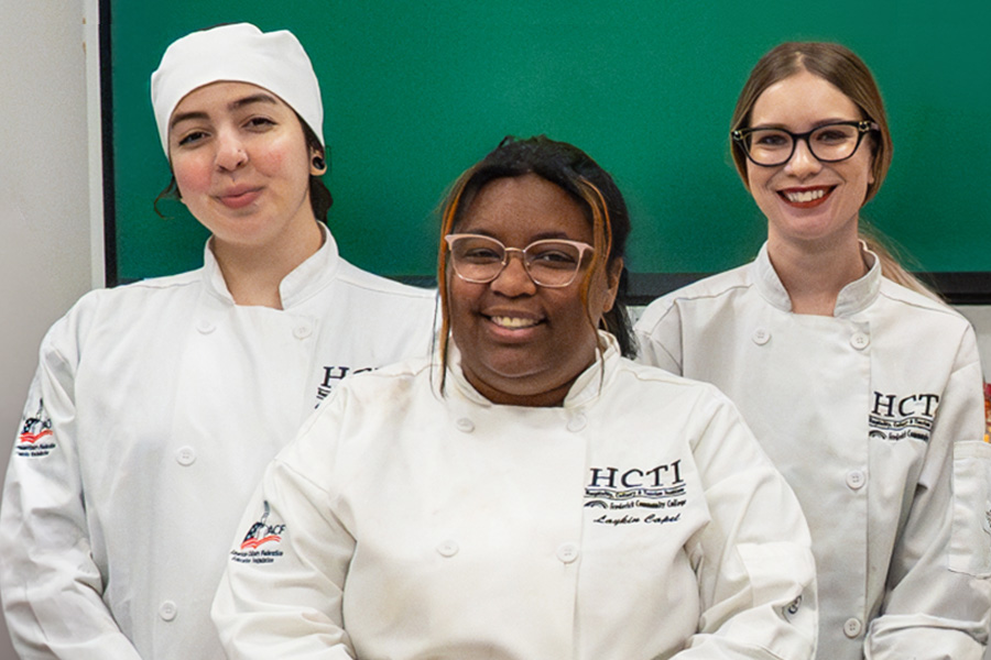 Three culinary baking & pastry HCTI students in white chef coats smile together in a classroom